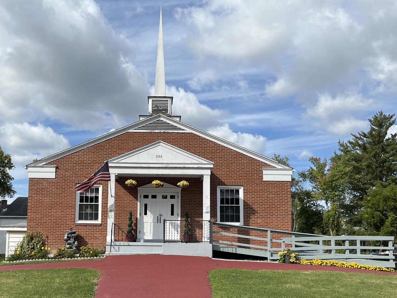 The front of First Baptist Church: a red brick building with a tall white steeple, a white columned entry with a flag beside the door, a red brick walkway across the lawn, and a ramp with a white railing at the right, under a blue sky with clouds.