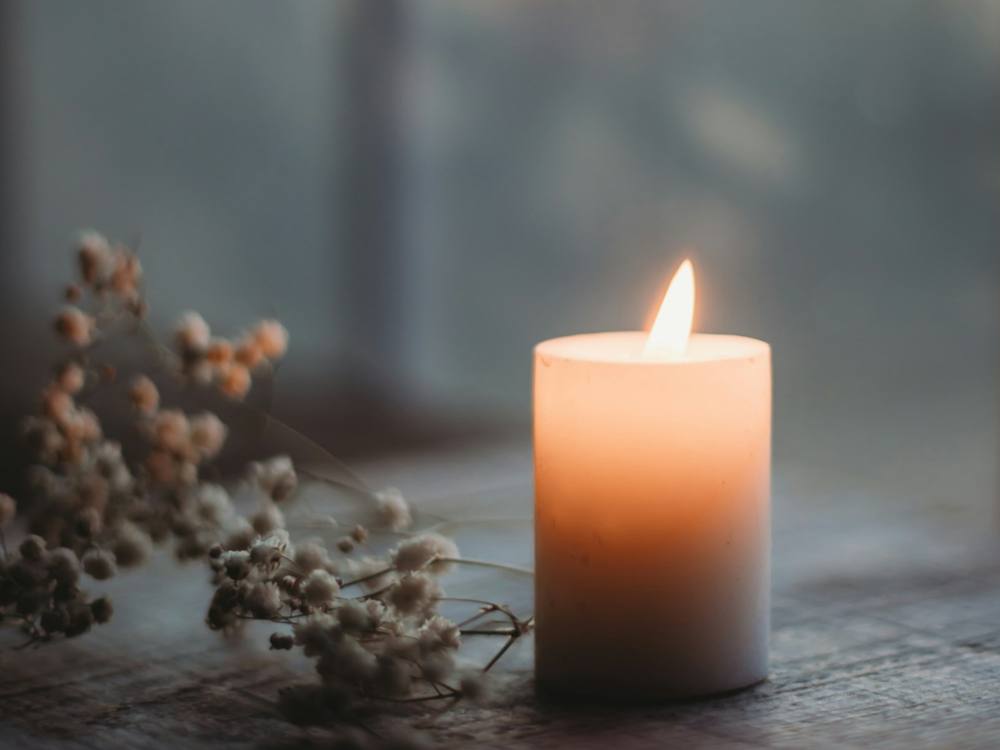 One lit candle in a short white holder on a wooden windowsill, with a sprig of dried flowers beside it, in soft window light.