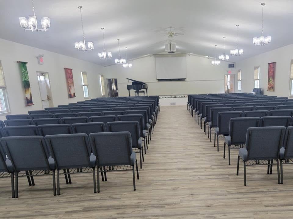 The sanctuary seen from the back: rows of gray padded chairs on both sides of a center aisle on a wood-look floor, with a piano, a screen, and banners at the front. No one is in the room.