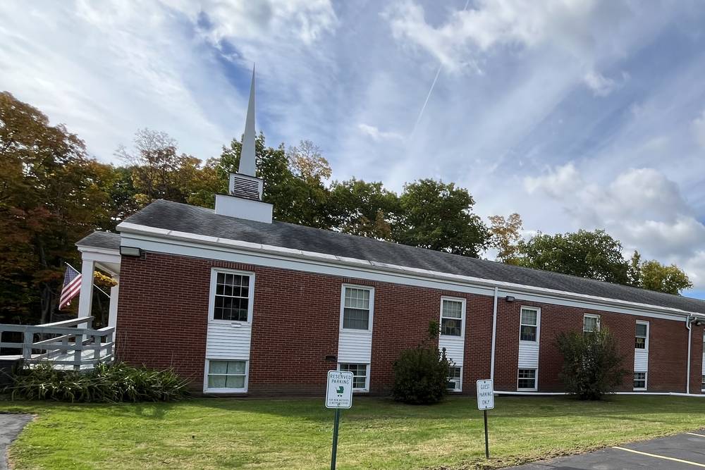 The long red brick side of the church and its steeple, beside a lawn and the edge of the parking lot, with two parking signs on posts.
